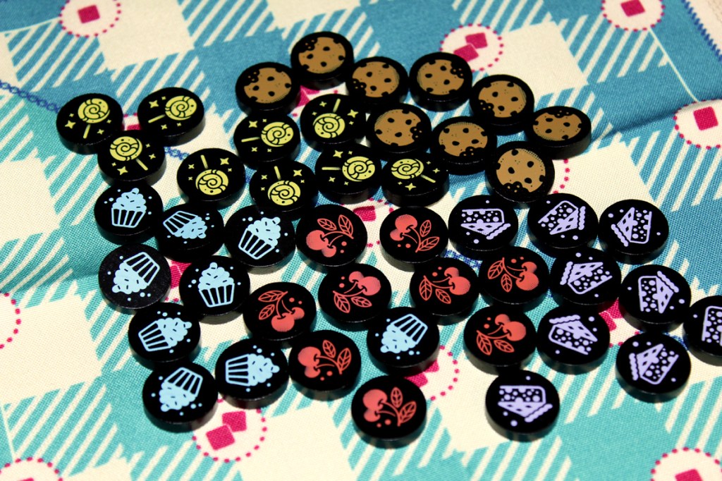 A collection of game tokens featuring various sweets, including cupcakes, cherries, and cookies, displayed on a patterned picnic blanket background.