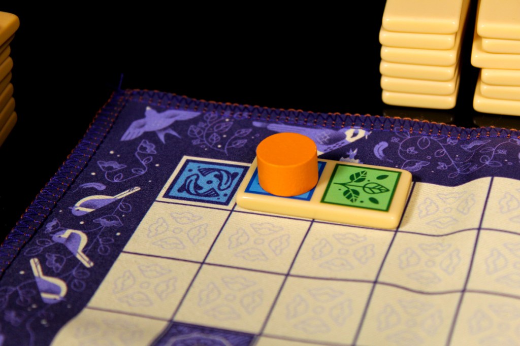 Close-up view of a game board featuring colored tiles and a player piece on a decorative fabric border.