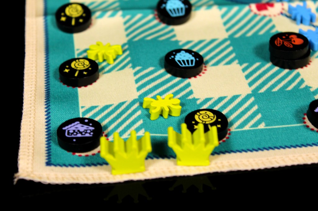 A close-up view of a colorful game board featuring various game tokens including sweets and ant meeples, set on a picnic-themed cloth.