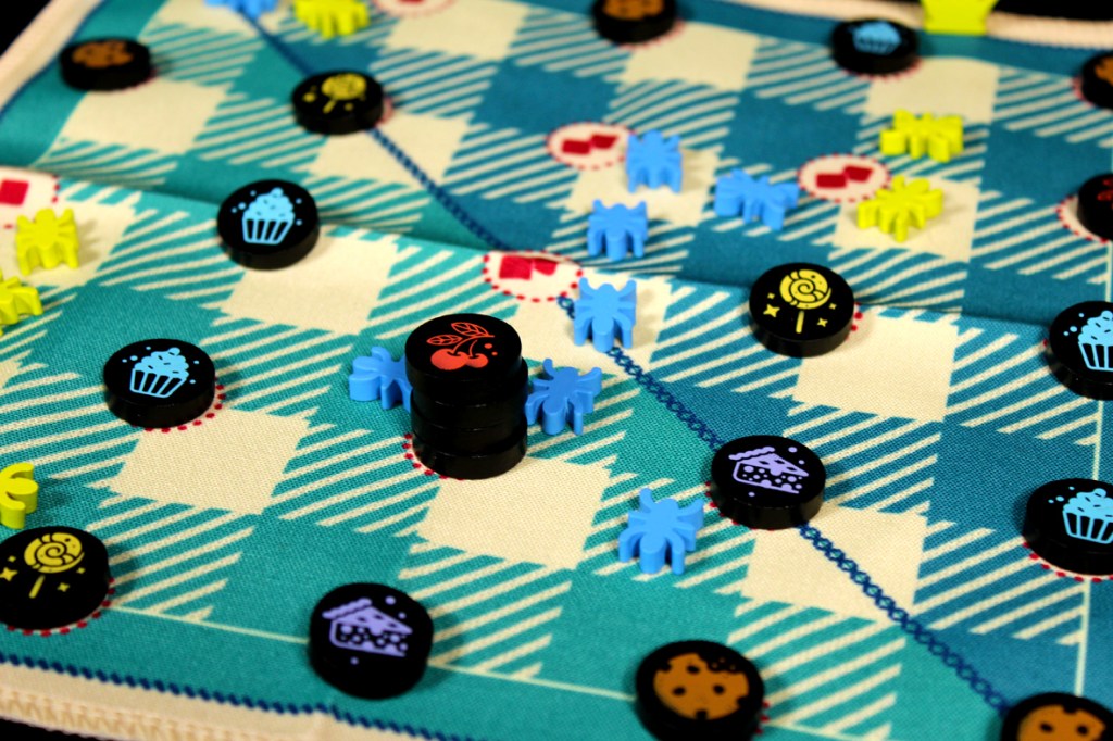 A close-up of a colorful picnic board game featuring various game pieces, including ants and food tokens on a patterned fabric board.