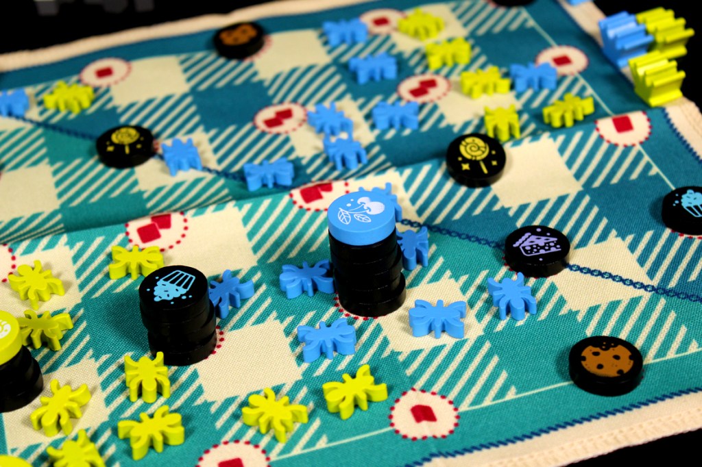 A colorful board game setup featuring blue, yellow, and black ant tokens on a patterned picnic blanket mat, with various food item tokens scattered across the board.