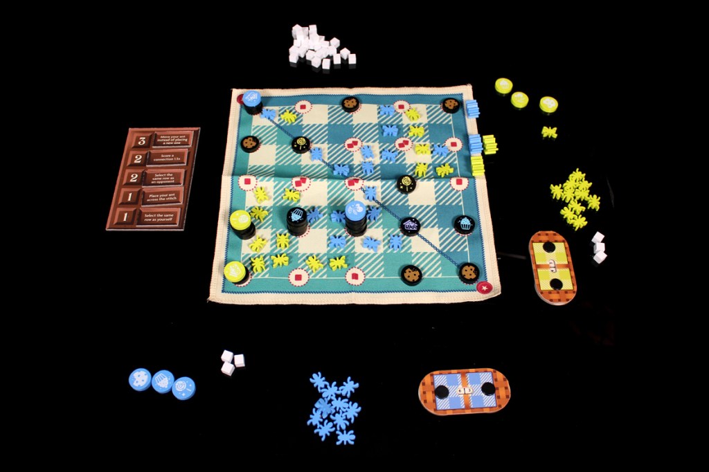 A tabletop game setup featuring a colorful game board with various tokens and pieces, including sugar cubes, cookie tokens, and player pieces, arranged on a black surface.