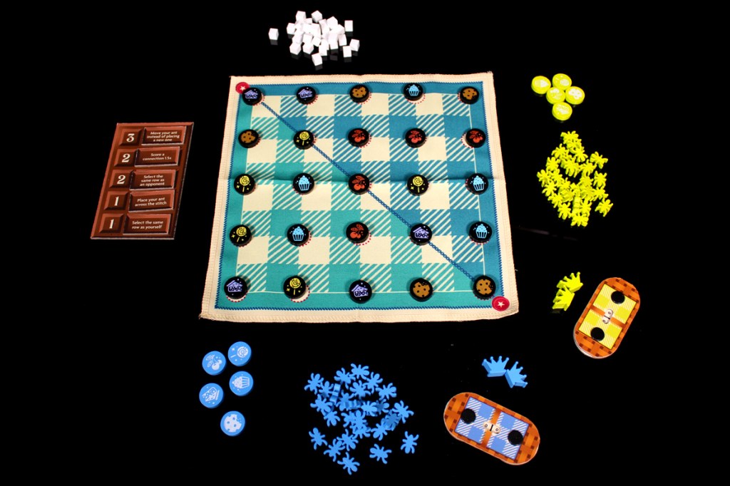 A colorful board game setup featuring a cloth game board with a checkered pattern, various tokens representing sweets, multiple player pieces shaped like ants, and game instruction cards arranged around the board.