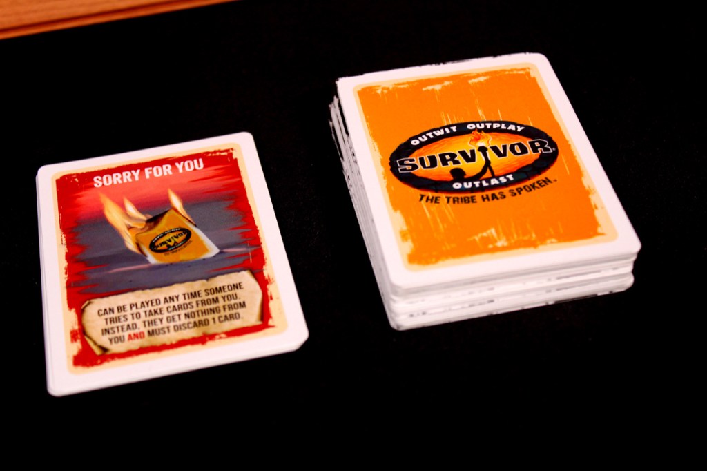 A board game card from Survivor: The Tribe Has Spoken, featuring a 'Sorry For You' card with flames and instructions on use, alongside a stack of game cards displaying the Survivor logo.