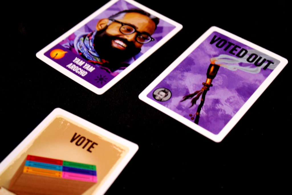 Three cards from the game Survivor: The Tribe Has Spoken, including a player character card with a smiling individual, a card labeled 'VOTED OUT', and a 'VOTE' card with colorful stacks.