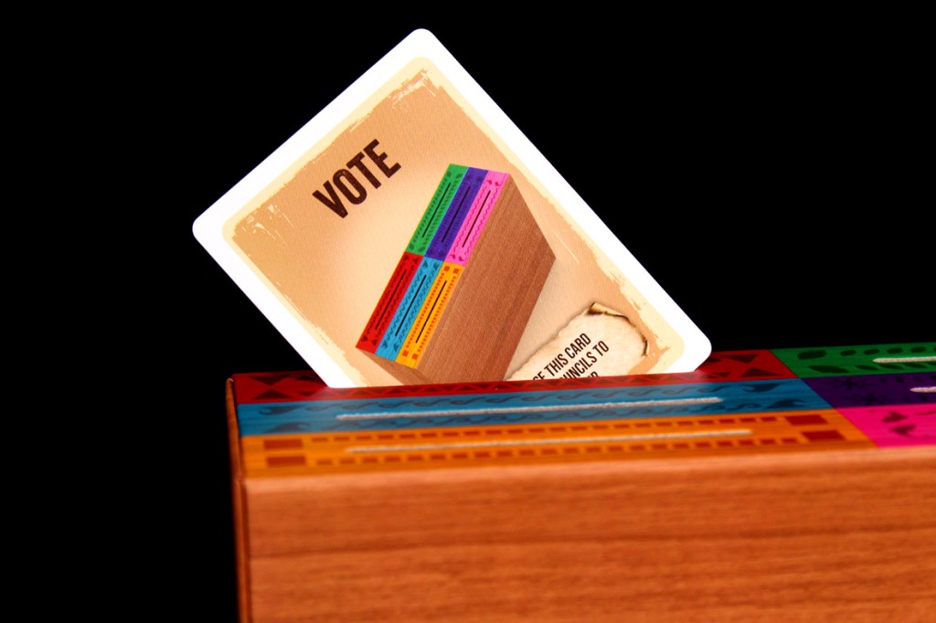 A 'Vote' card partially inserted in a colorful board game box, featuring various tribal designs.