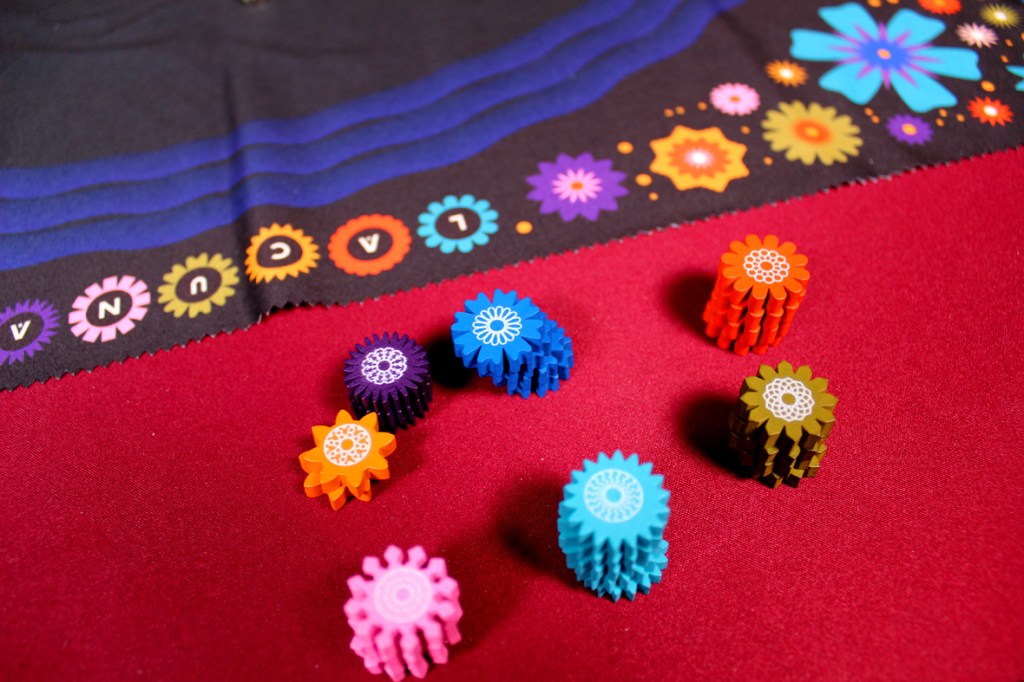 Colorful flower tokens on a game mat with the title 'Lacuna' at the edge, resting on a red surface.