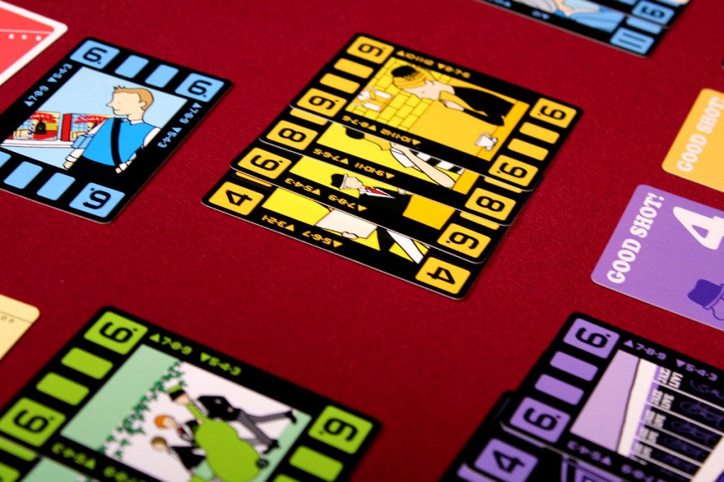 A close-up of colorful game cards laid out on a red table, with illustrations of characters and indicators such as 'Good Shot!' showcasing game elements from 'Wind the Film'.