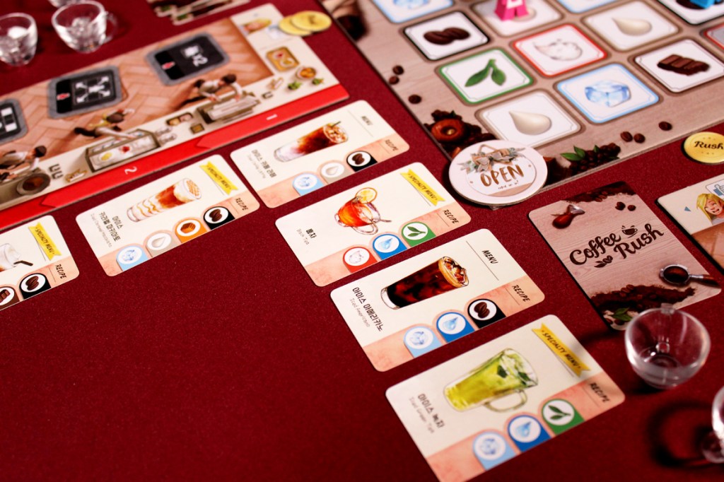 An overhead view of a board game setup for 'Coffee Rush', featuring player menu cards, a game board with icons for ingredients and orders, along with small cups and tokens on a burgundy background.