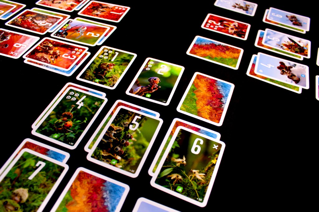 A vibrant tabletop setup of the card game Pixies, featuring colorful cards with numbers and illustrations of small creatures and nature, arranged on a black surface.