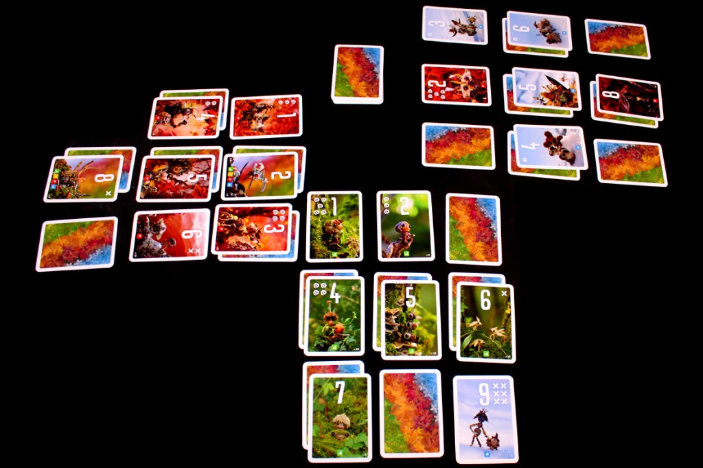 A tabletop game setup featuring colorful cards spread out on a black surface. The cards display various characters and numbers, indicating gameplay elements of the game 'Pixies'.