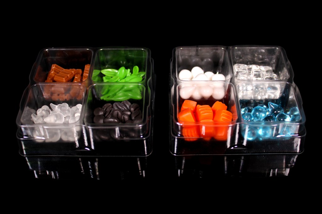A clear tray containing various colored game pieces for a coffee-themed board game, including brown, green, black, white, orange, and blue pieces, arranged in compartments.