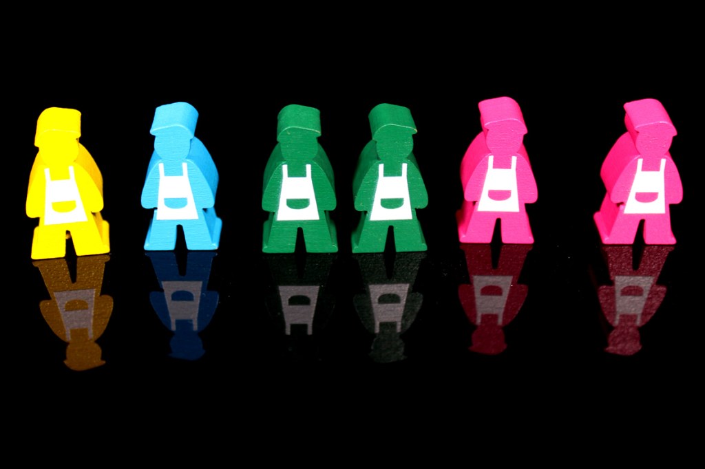 Colorful wooden meeples representing baristas from the game Coffee Rush, arranged in a row against a reflective black background.