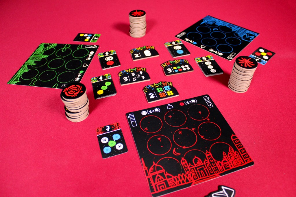 A colorful board game setup featuring various tiles, tokens, and objective cards, all themed around a fireworks celebration, displayed on a red background.