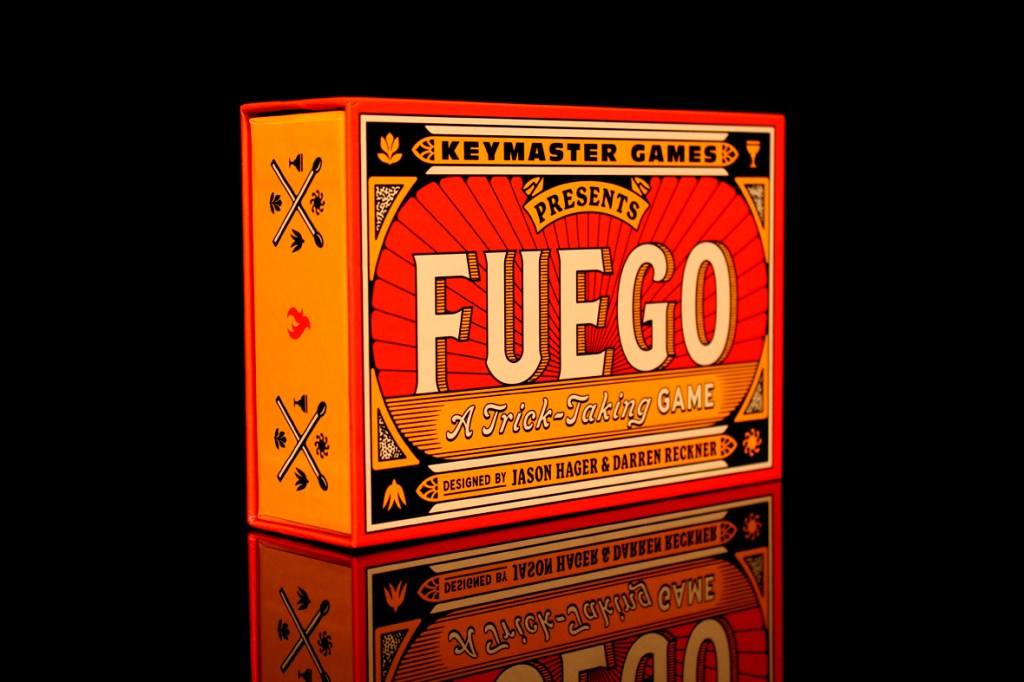A brightly colored box of the board game 'Fuego' with the text 'Presents Fuego: A Trick-Taking Game' on the front, surrounded by fire-themed illustrations, reflecting on a black surface.