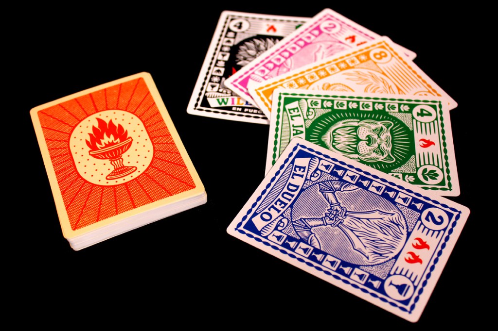 A deck of playing cards featuring a fiery motif with a lit torch design on the top card, surrounded by various colored cards displaying different values and symbols.
