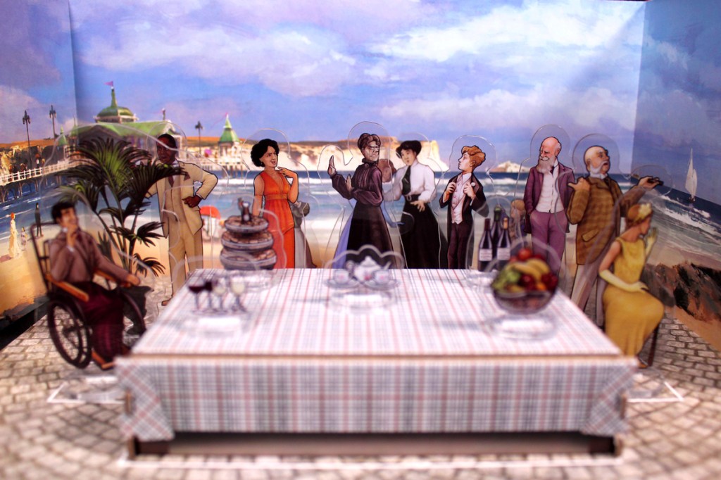 A colorful tabletop setup for the board game Picture Perfect, featuring various character pieces arranged around a decorated dining table with a seaside backdrop.