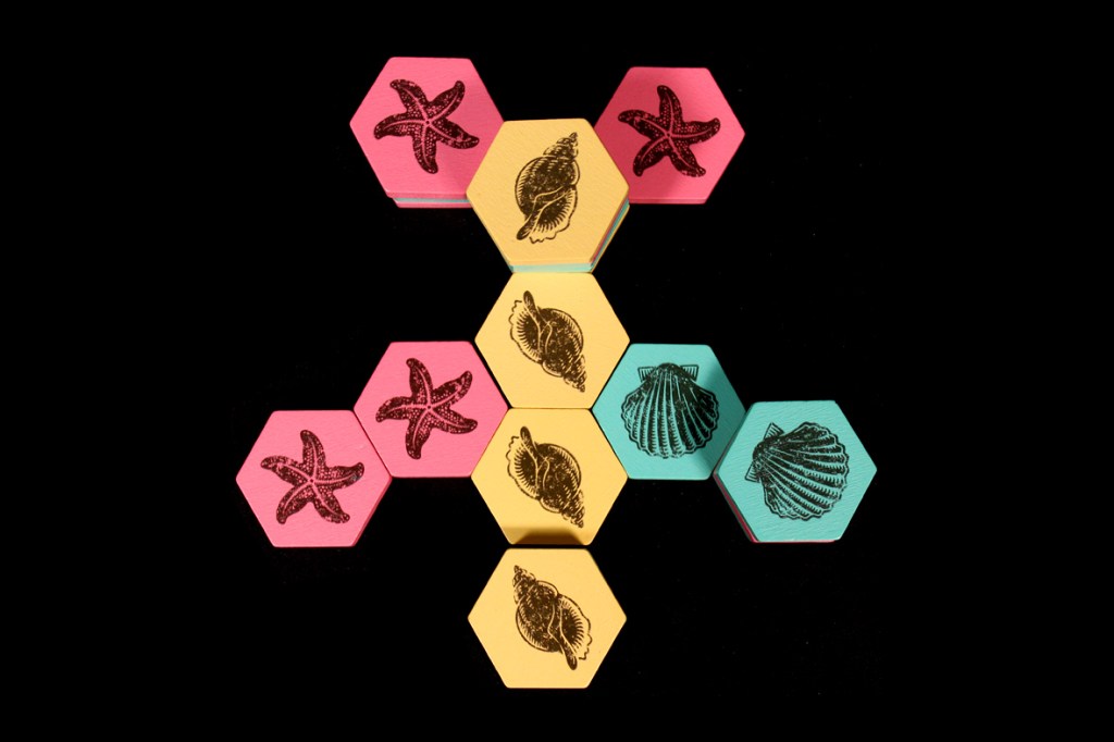 A collection of colorful hexagonal game tiles in pink, yellow, and teal, featuring engravings of starfish and seashells, arranged in a central formation.
