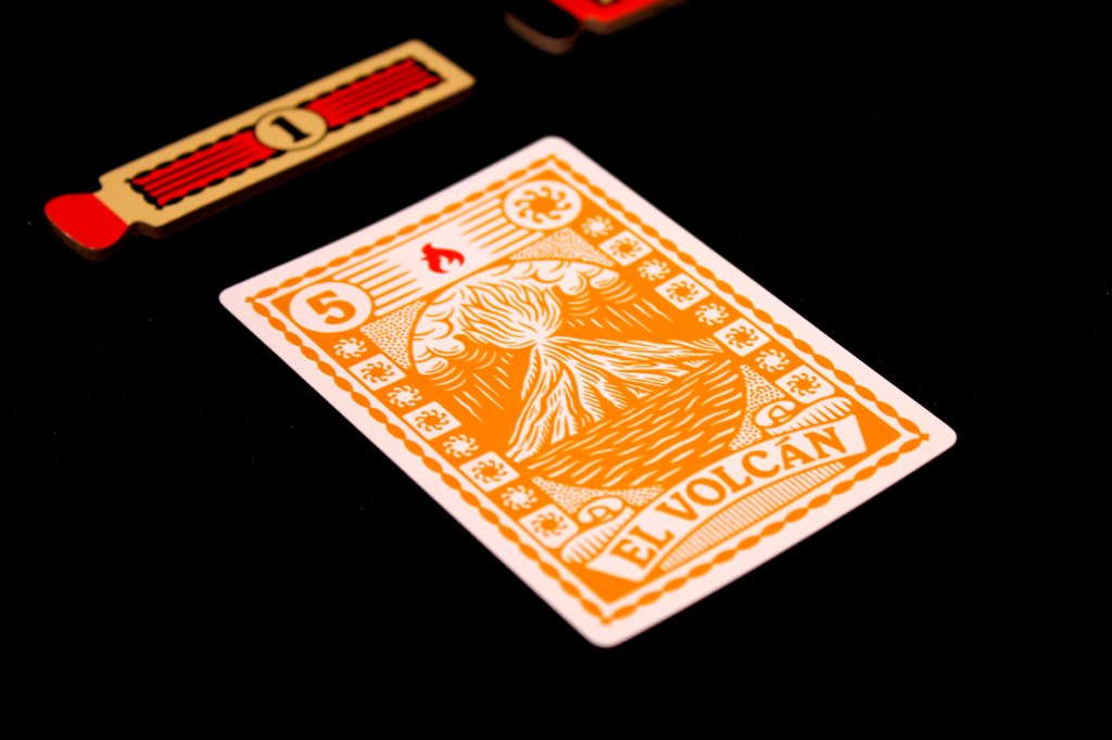 An orange card from the board game Fuego featuring an illustration of a volcano and the text 'El Volcán' with a value of 5, placed next to a red Strike Token.
