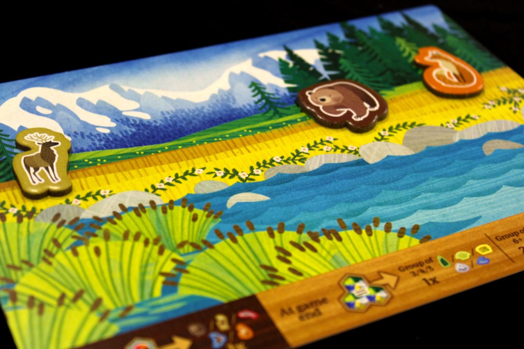 Close-up of a gameplay board for Cascadia Junior featuring a colorful landscape with animals like a moose, bear, and fox on a riverbank.