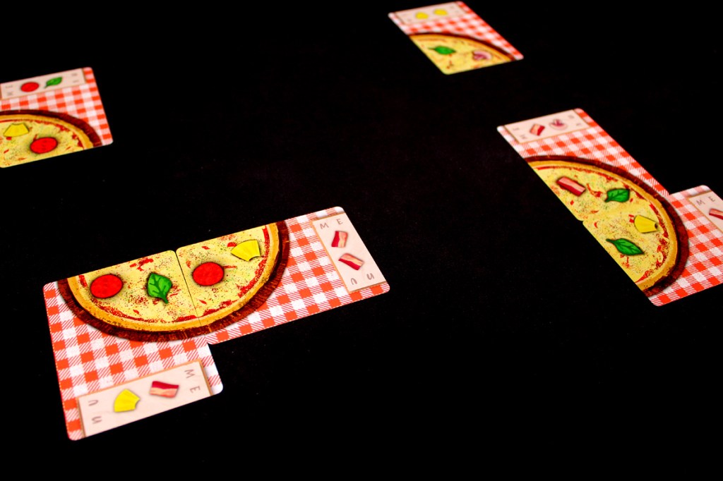 Game cards depicting pizzas with various toppings, laid out on a checkered tablecloth background.