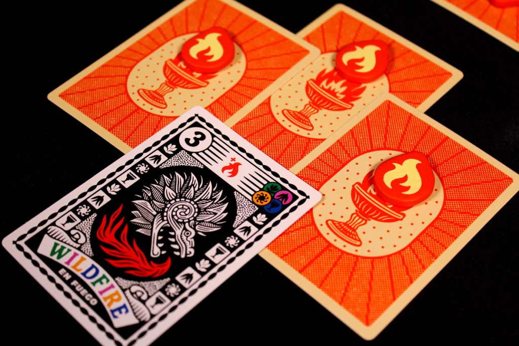 A Wildfire card from the game Fuego, featuring a vibrant design with a stylized flame and decorative elements, placed in front of three Flame Tokens on matching orange background cards.