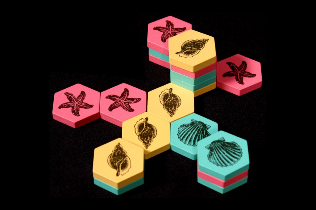 Colorful hexagonal game tiles displayed in a stack, with designs of sea creatures like starfish and shells, set against a dark background.