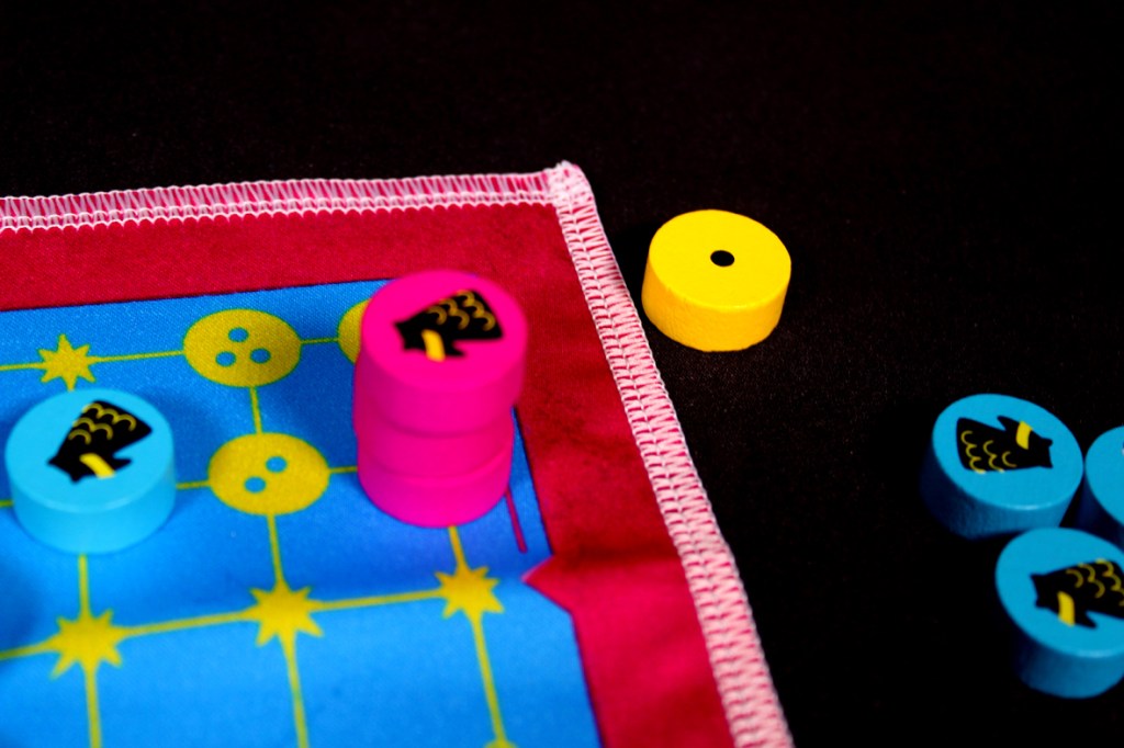 Close-up of a colorful game board with various wooden tokens in pink, blue, and yellow, featuring a blue sky background with geometric patterns.