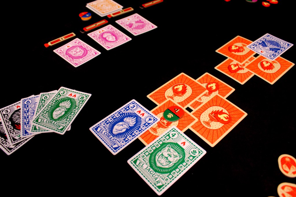 A tabletop setup for the game Fuego, featuring colorful playing cards with various symbols and tokens arranged on a black surface.