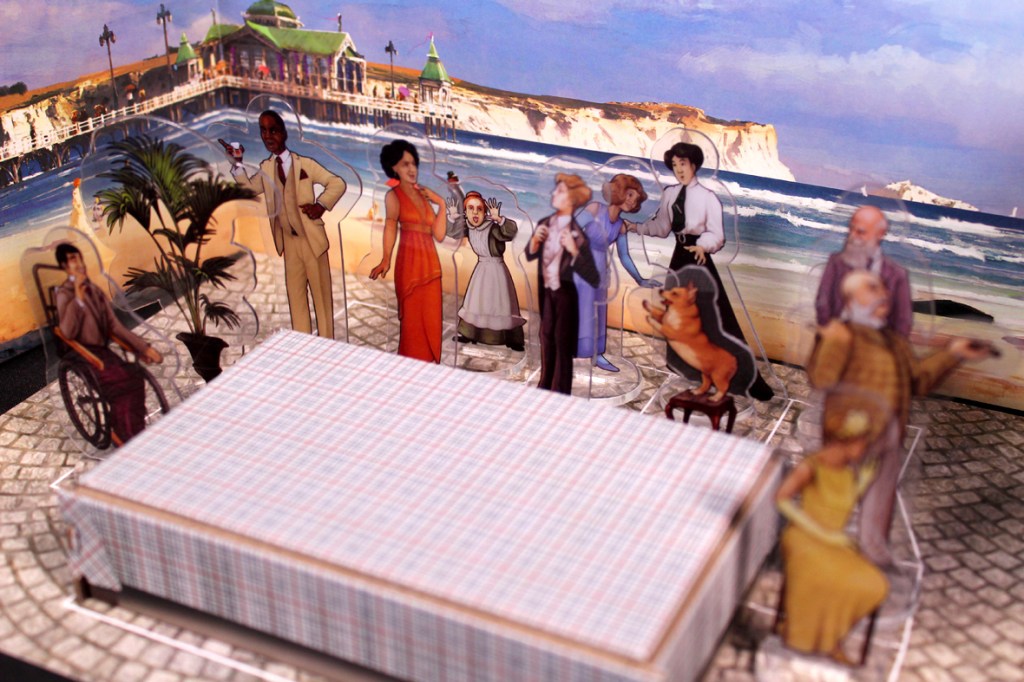 A tabletop game setup for 'Picture Perfect' featuring various character cutouts arranged around a plaid table, with a seaside background.