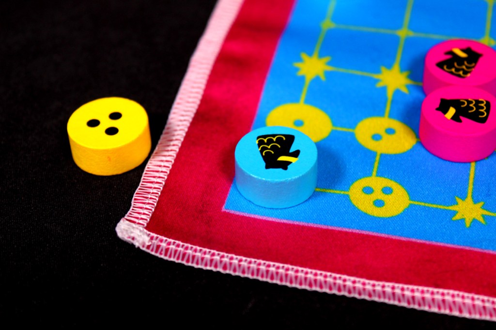 Close-up of game pieces on a colorful board. A yellow piece with two black dots is positioned next to a light blue piece with a black symbol, alongside other pink and blue pieces on a blue game mat.