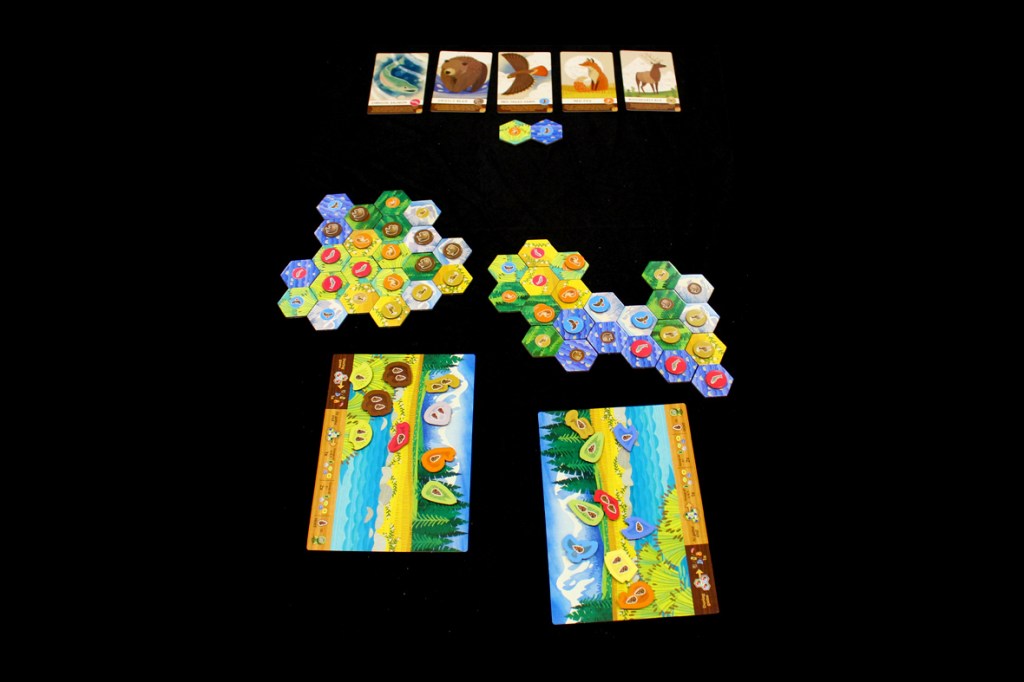A layout of the board game Cascadia Junior featuring hexagonal tiles representing different habitats and wildlife, along with player boards depicting a scenic panorama.