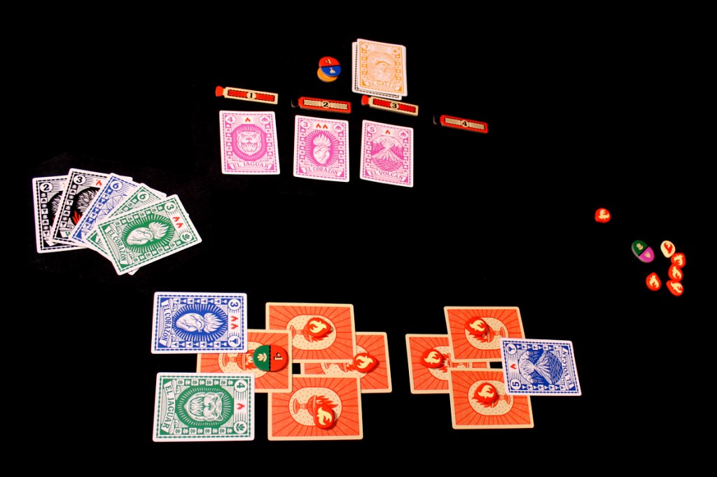 A tabletop game setup displaying multiple playing cards in various colors (blue, green, black, pink) along with flame tokens and score trackers on a black background.