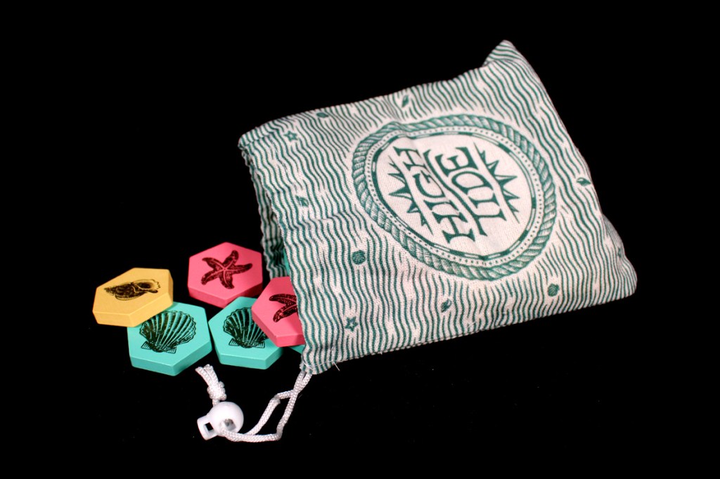 A fabric bag labeled 'High Tide' with a wavy pattern, alongside colorful hexagonal game tiles in yellow, pink, and teal featuring ocean-themed illustrations.