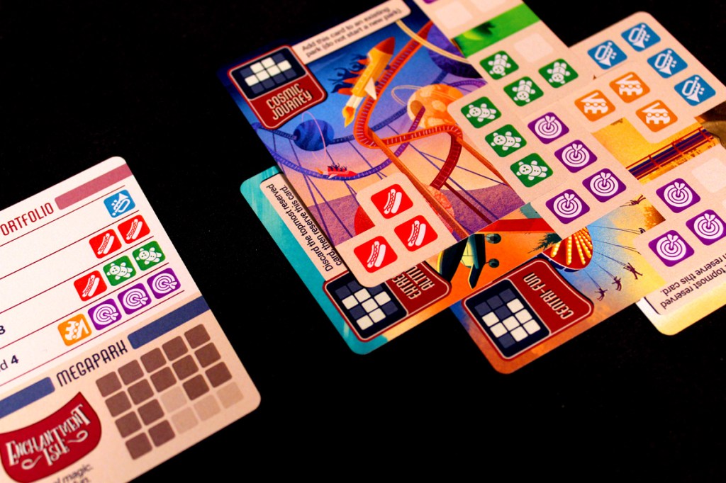 An overhead view of game components for 'Lands of Amazement', showing various cards including Portfolio and Megapark cards, alongside colorful themed cards depicting attractions.