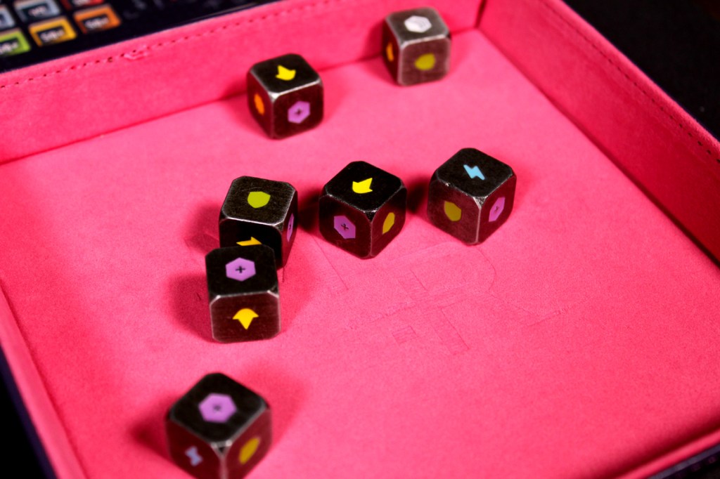 A close-up view of several colorful dice with various symbols, resting in a pink felt-lined box, likely associated with a board game.