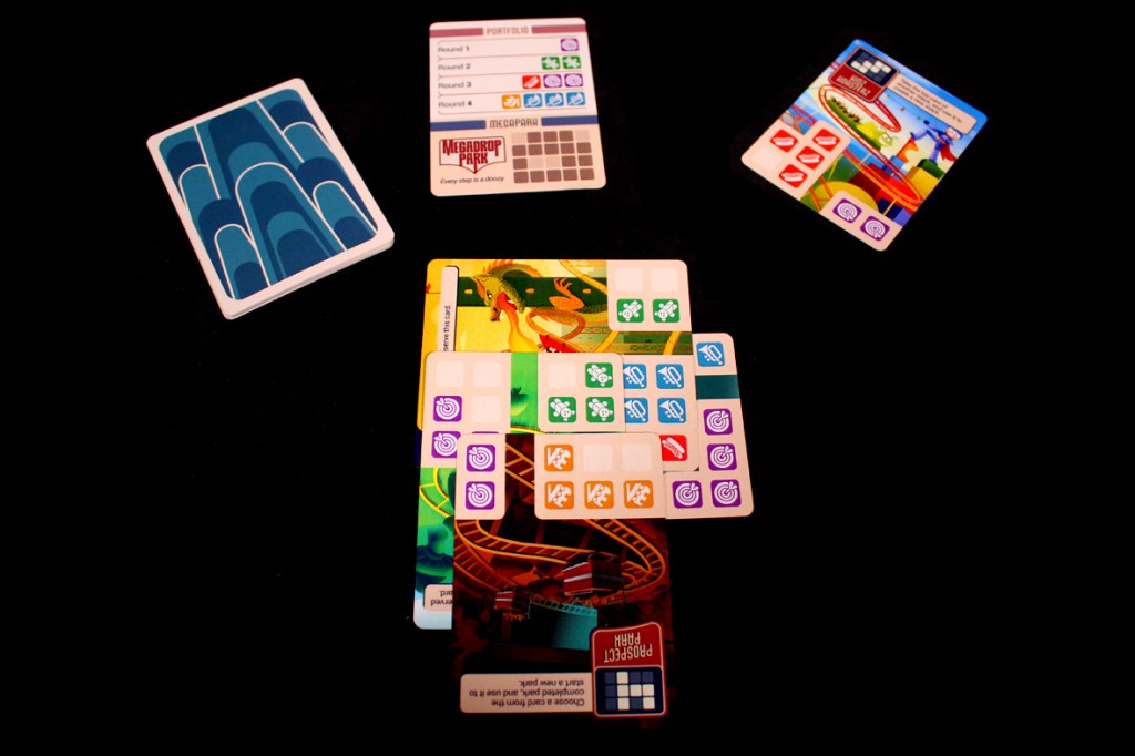 Image of various game cards from the board game 'Lands of Amazement', including a Megapark card, Portfolio card, and several park cards with colorful designs and icons on a dark background.