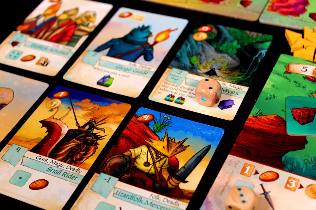 A close-up of game cards from 'Creature Caravan', featuring illustrations of characters like a giant snail rider, a lizardfolk mercenary, and other fantasy elements, alongside dice and game tokens.
