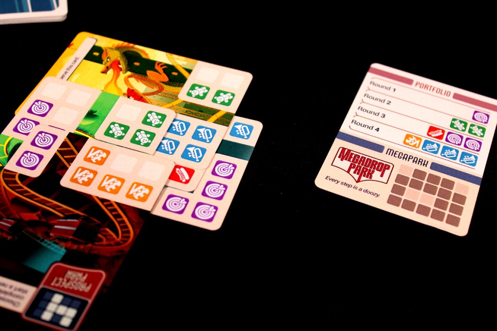 Close-up of game cards and a portfolio sheet from the board game 'Lands of Amazement,' featuring colorful icons and shapes for gameplay mechanics.