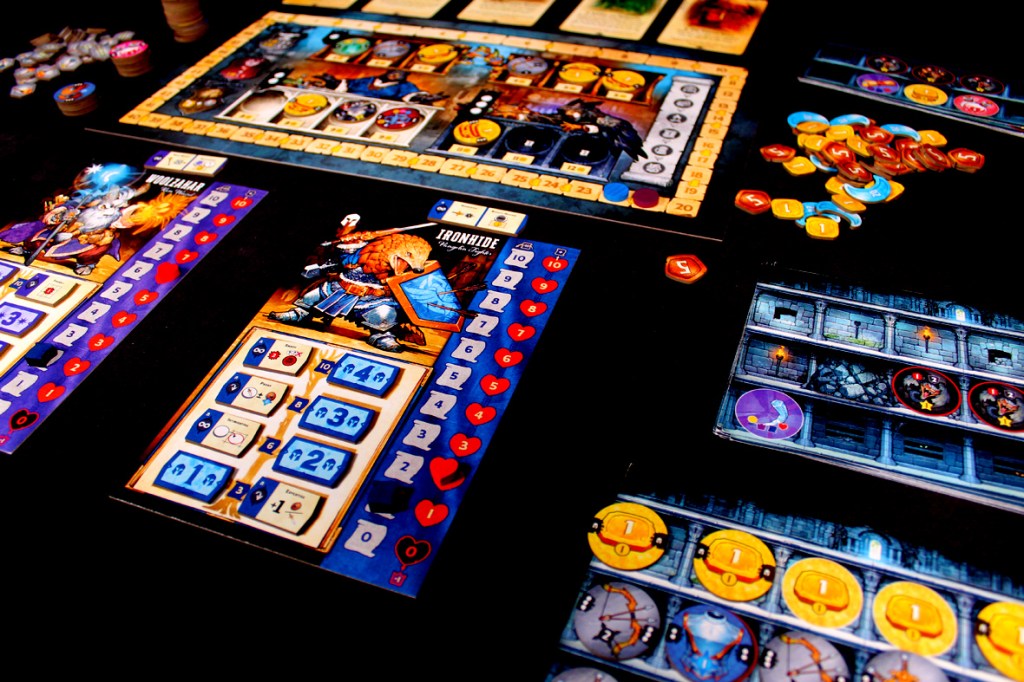 Close-up of a tabletop board game featuring player boards with character illustrations, health and resource trackers, and various game tokens and components spread out on a black surface.