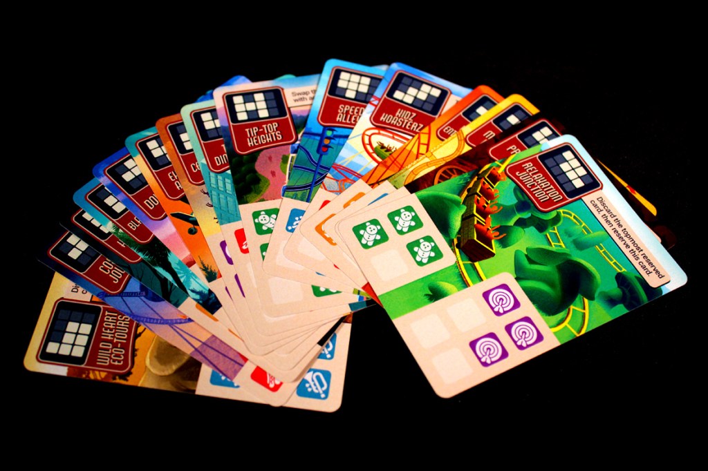 A spread of colorful game cards from the board game 'Lands of Amazement', featuring various park attractions and gameplay elements, displayed against a black background.