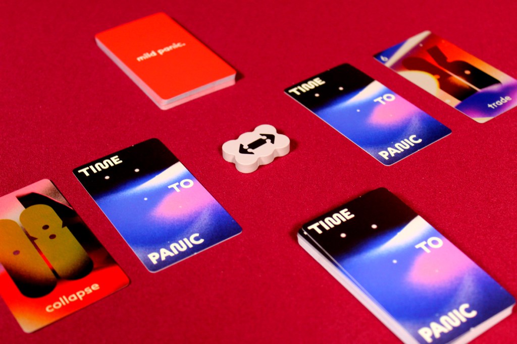 A flat lay image featuring several game cards from the board game 'Time to Panic' on a red background, including 'collapse' and 'trade' cards, with a panic card deck visible above and a gap token in the center.