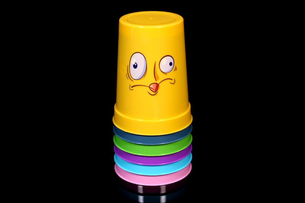 A stack of colorful plastic cups with a cartoonish face on the top yellow cup, featuring large eyes and a surprised expression, set against a black background.
