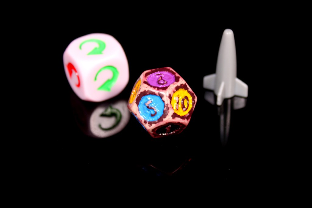 A pink die with numbers 2, 3, and a green question mark, a multi-colored die showing numbers 5 and 10, and a gray rocket-shaped piece, all against a black background.