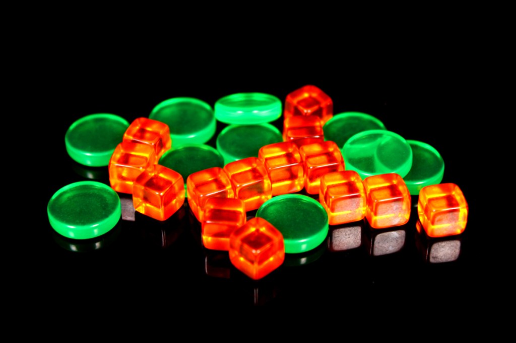 A collection of green and orange game tokens, including circular and cube shapes, arranged on a reflective black surface.
