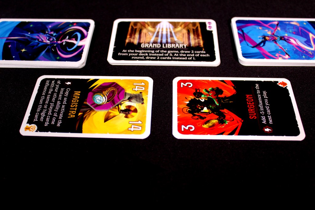 A flat lay of several game cards featuring colorful artwork. The foreground includes two prominent cards labeled 'MAGISTRA' and 'SURGEON,' while two additional cards and one larger card labeled 'GRAND LIBRARY' are positioned in the background.