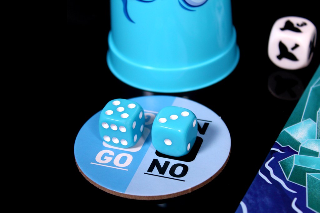 A blue cup, two dice on a 'GO/NO' game space, and a seal die are displayed on a dark surface, related to the game Push Push Penguin.