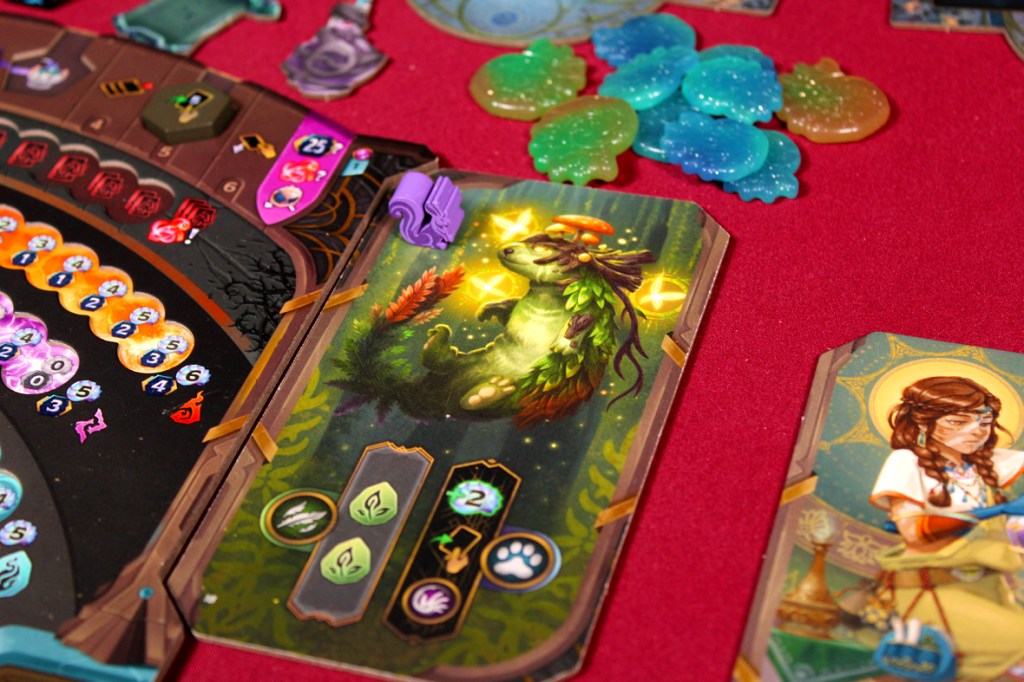 A close-up of a board game setup featuring colorful tokens, potion cards, and player boards with various stats and artwork. The table is covered in a red cloth, highlighting the intricate game design.