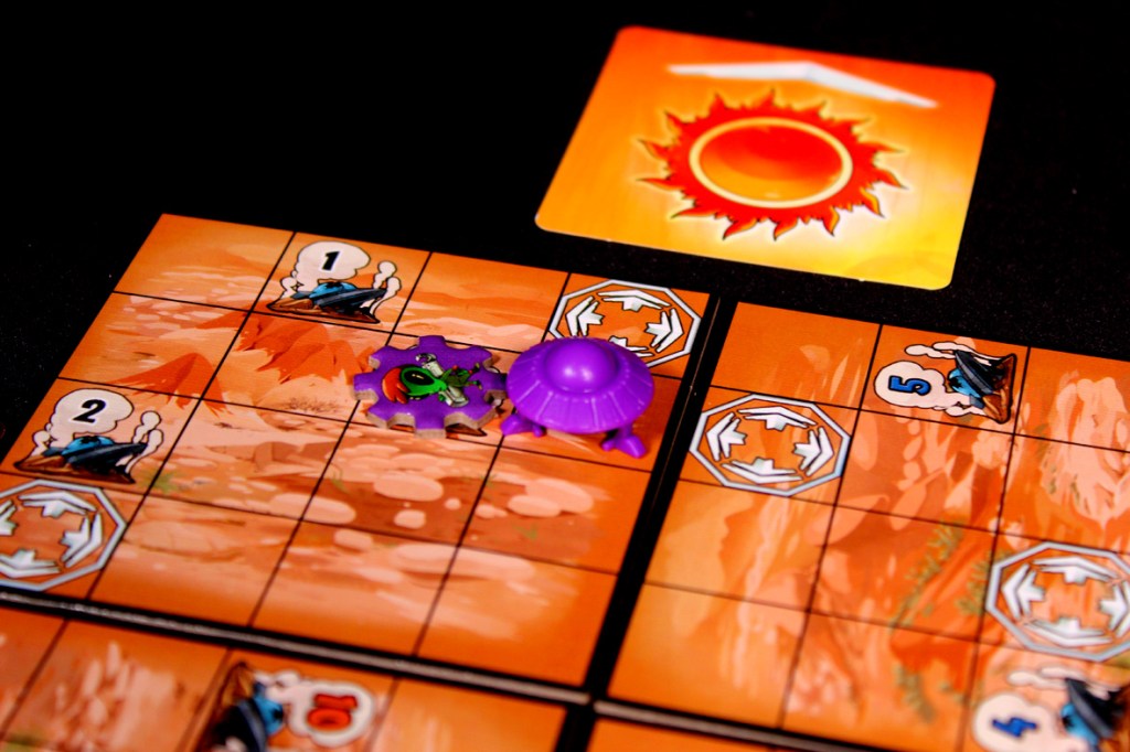 A board game setup featuring a bright orange game board with numbered spaces and movement markers. A purple spaceship and an alien figure are positioned on the board, along with a visible sun card.