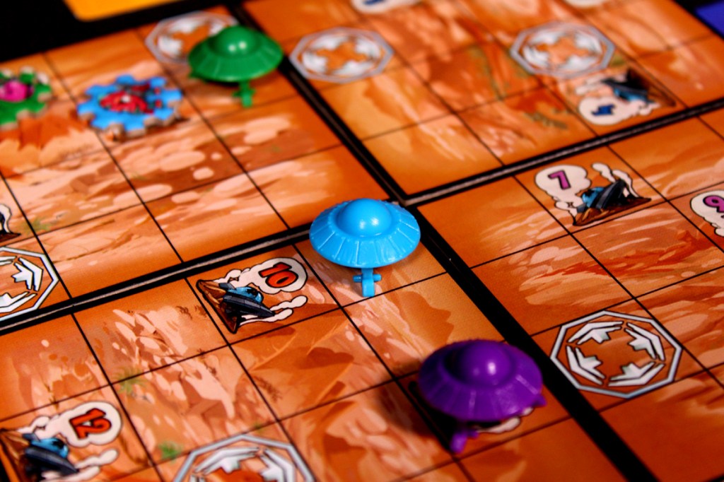 Close-up shot of a board game setup for 'Crash & Grab', showing colorful saucer pieces (blue, green, and purple) on a vibrant game board with numbered spaces.